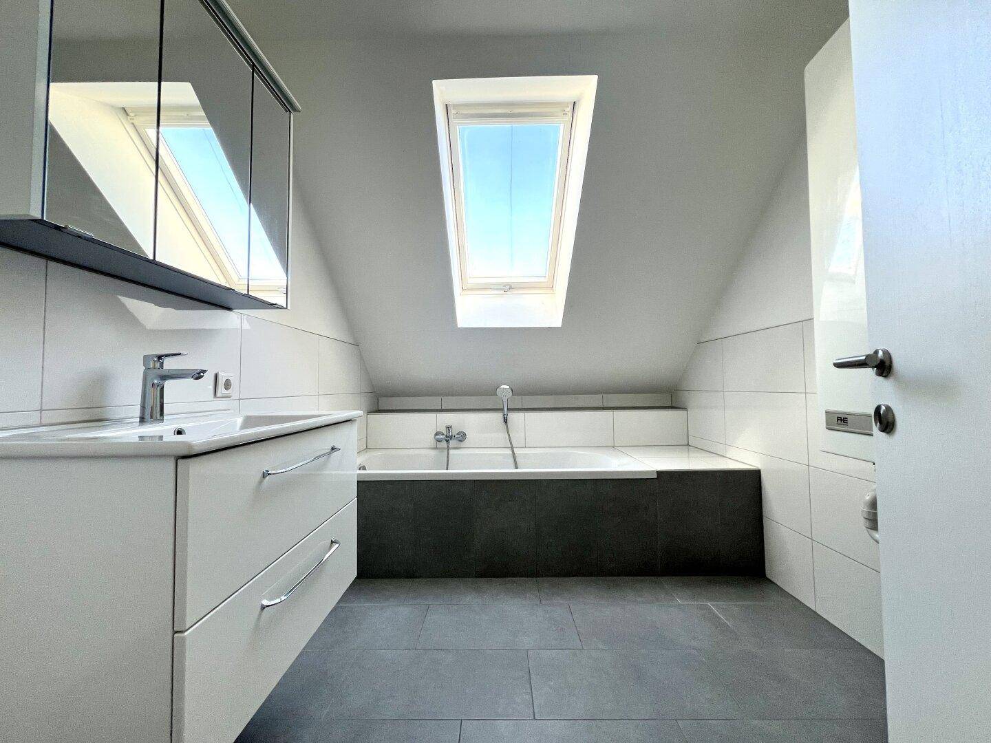 Modernes Badezimmer im Dachgeschoss mit Badewanne, Waschtisch, Spiegelschrank und Dachfenster.