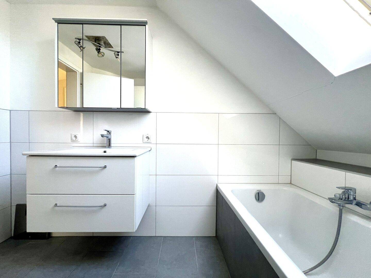 Badezimmer mit Waschtisch, Spiegelschrank und Badewanne unter einem Dachfenster.
