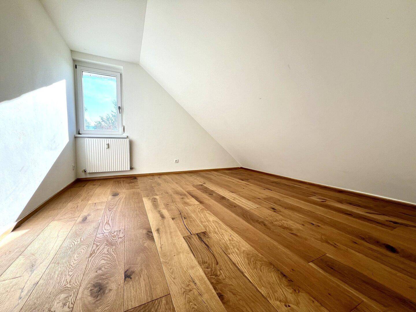 Helles Schlafzimmer im Dachgeschoss mit Holzboden, schräger Decke und Fenster mit Rollladen.