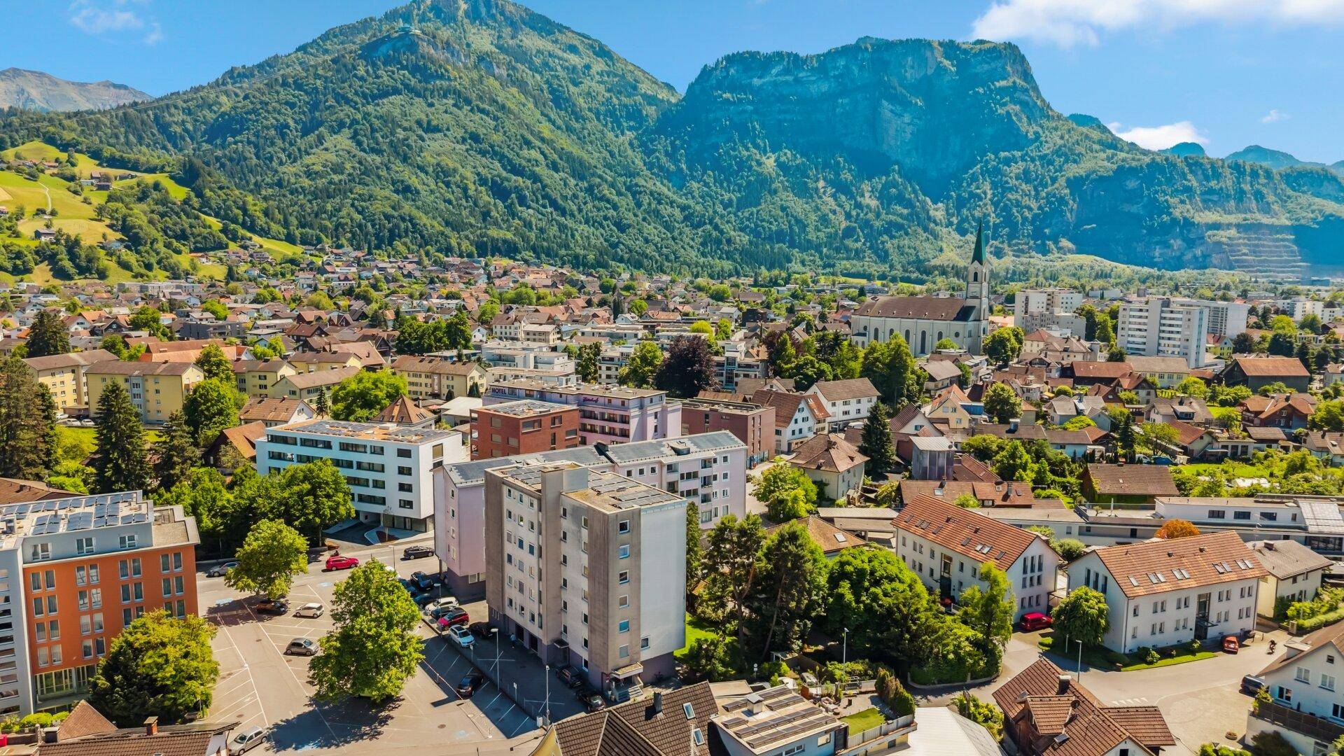Panoramablick auf eine Stadt mit Wohngebäuden und Bergen im Hintergrund unter blauem Himmel.