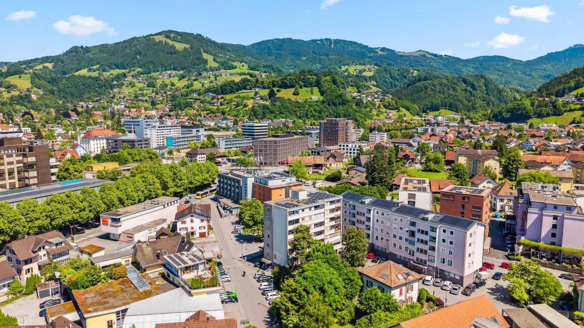 Weitläufiger Blick über eine Stadt mit Wohngebäuden, grünen Hügeln und klarem Himmel.