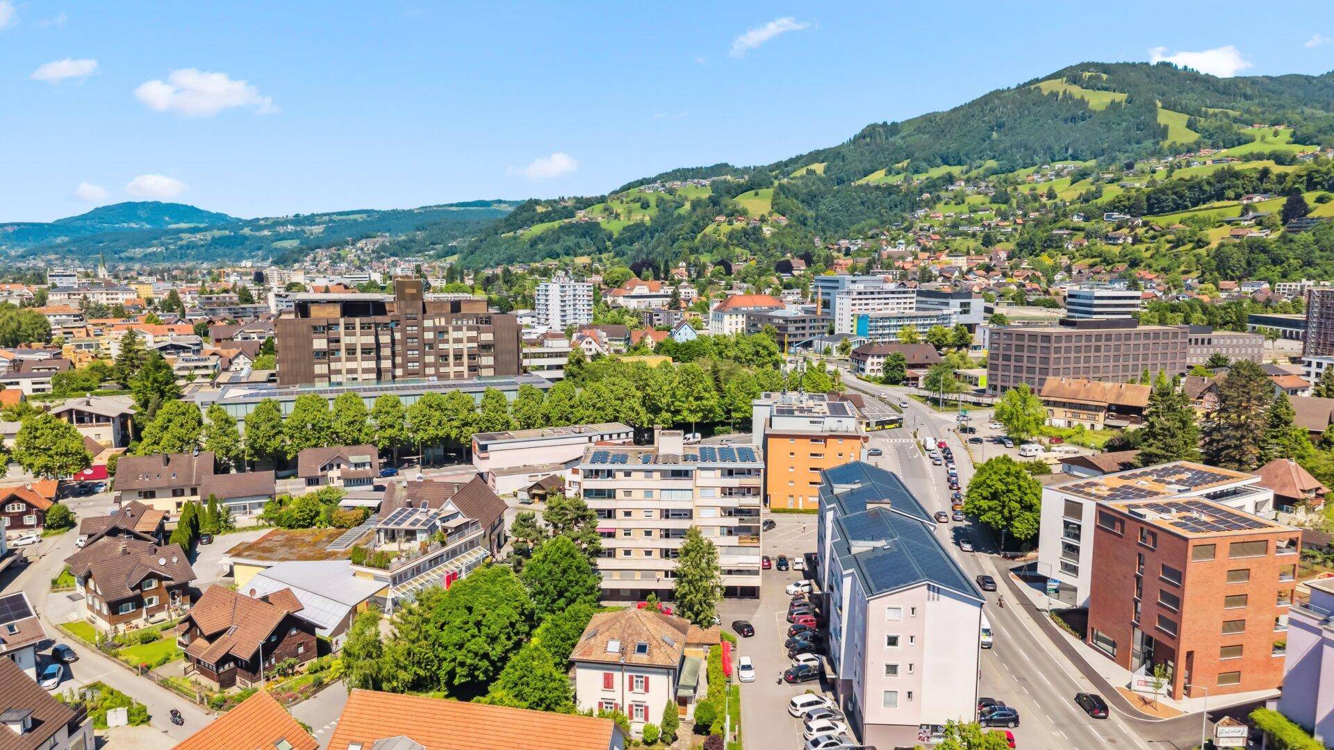 Luftaufnahme einer Stadt mit Wohn- und Geschäftsgebäuden entlang einer Hauptstraße und grünen Hügeln.