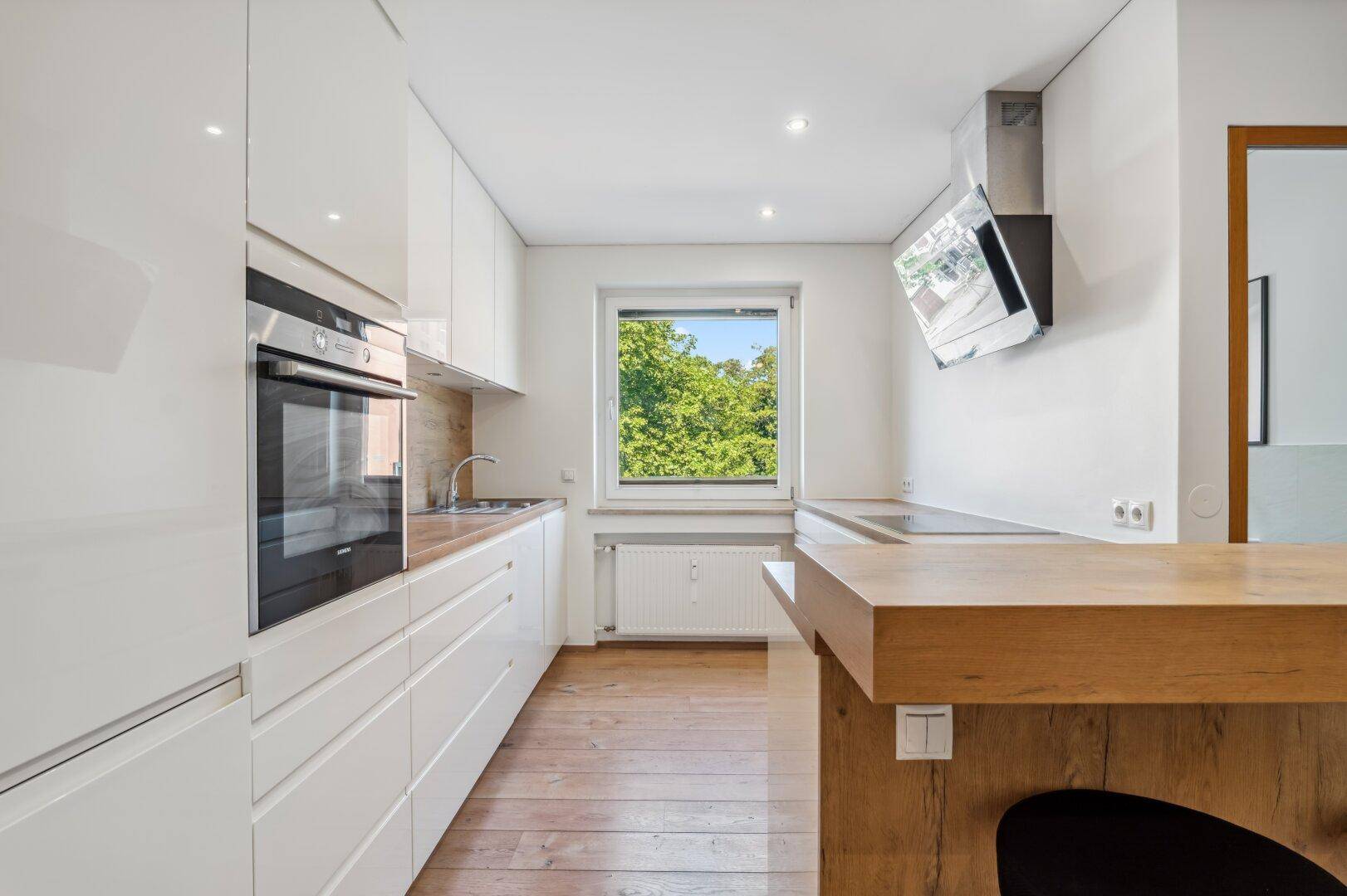 Moderne Küche mit weißen Hochglanzfronten, Holzarbeitsplatte und Fensterblick.