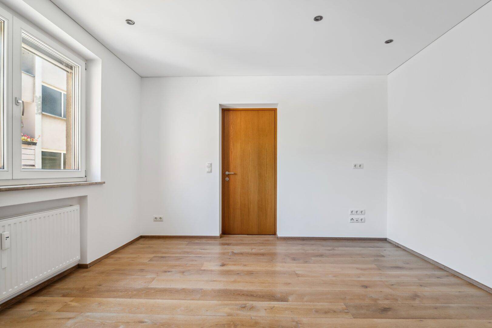 Leeres Schlafzimmer mit Holzboden, weißen Wänden und Fenster.