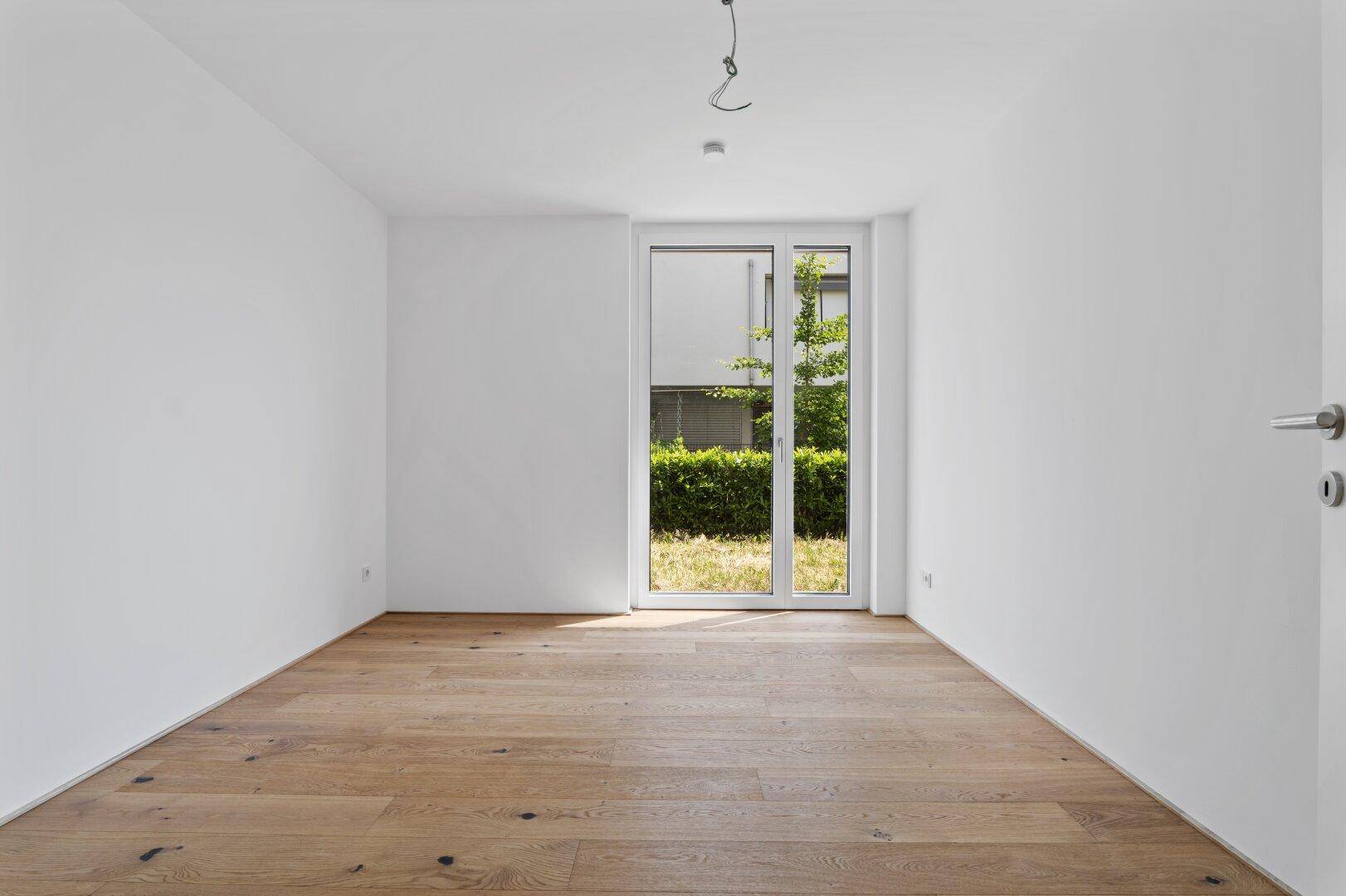 Helles Zimmer mit Holzboden und großen Fenstern mit Blick ins Grüne.