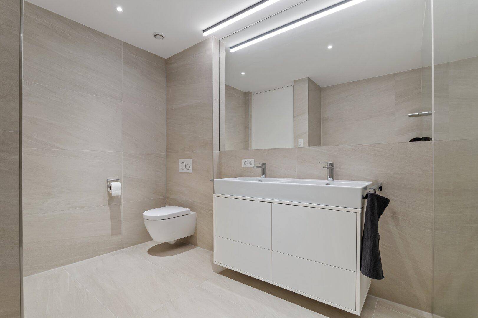 Geräumiges Badezimmer mit Doppelwaschbecken, Wand-WC und begehbarer Dusche.