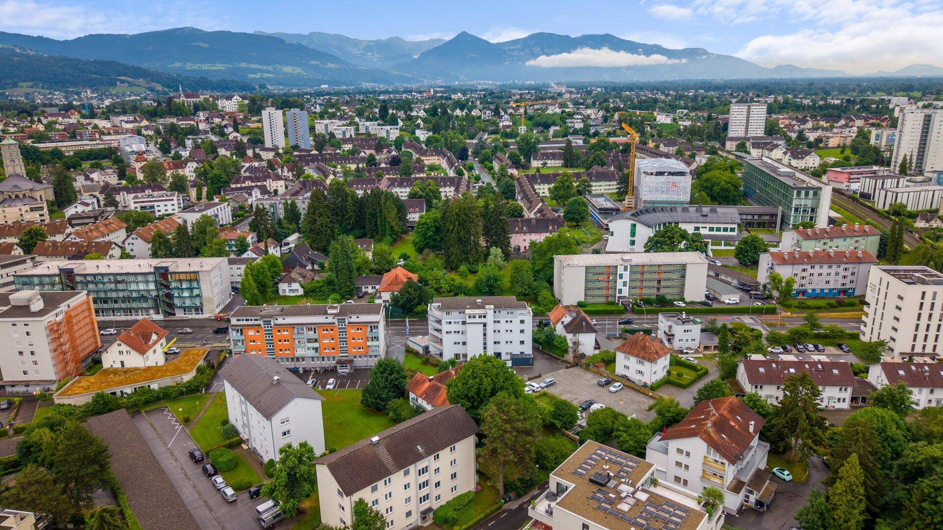 Weitwinkel-Luftaufnahme einer Stadtlandschaft mit Bergen im Hintergrund unter einem blauen Himmel.