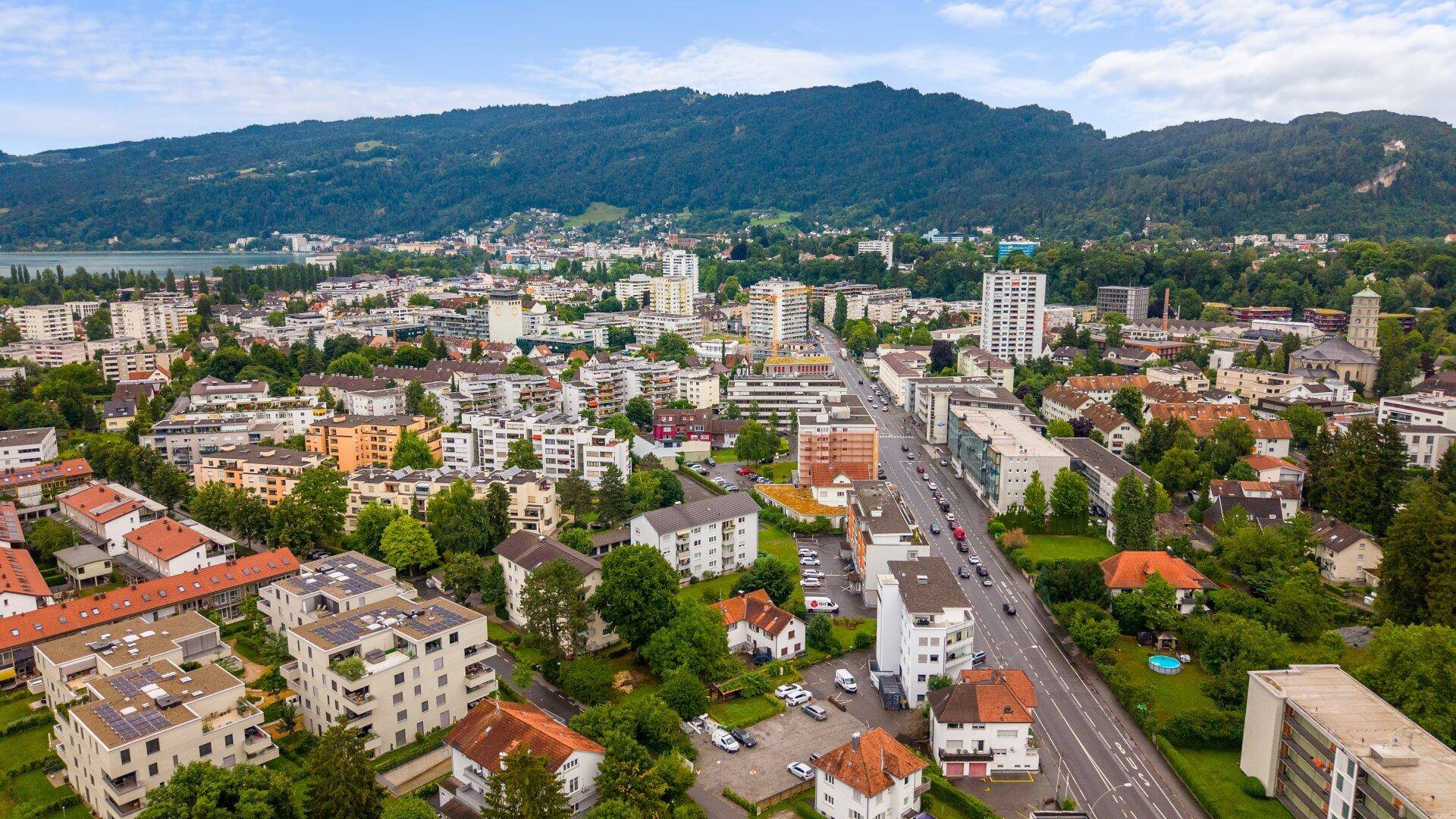 Luftaufnahme einer Stadt am Seeufer mit bewaldeten Hügeln und Bergen im Hintergrund unter blauem Himmel.