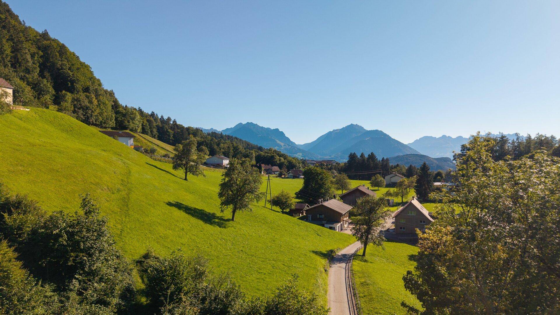 Panoramablick auf ein Tal mit Häusern, einer Straße, grünen Hügeln und fernen Bergen.