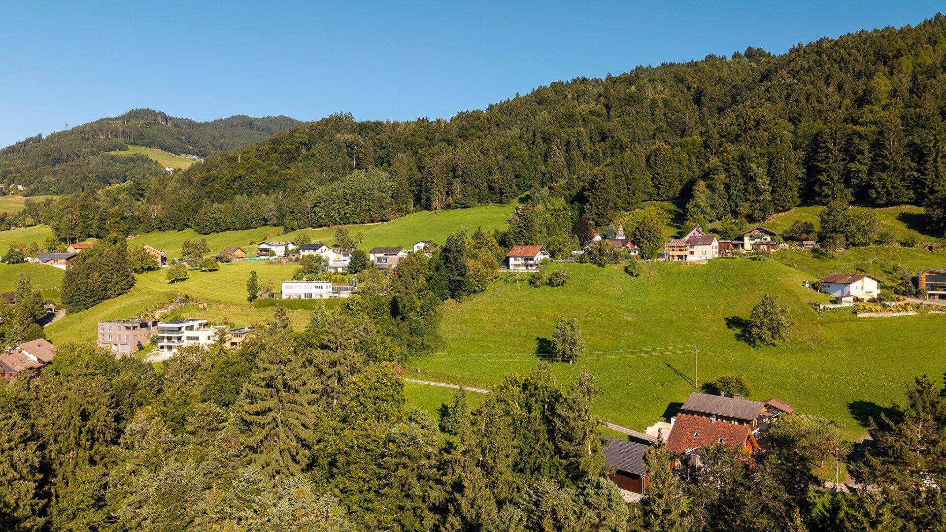 Luftaufnahme eines malerischen Dorfes eingebettet in eine grüne, hügelige Landschaft mit Bergen.