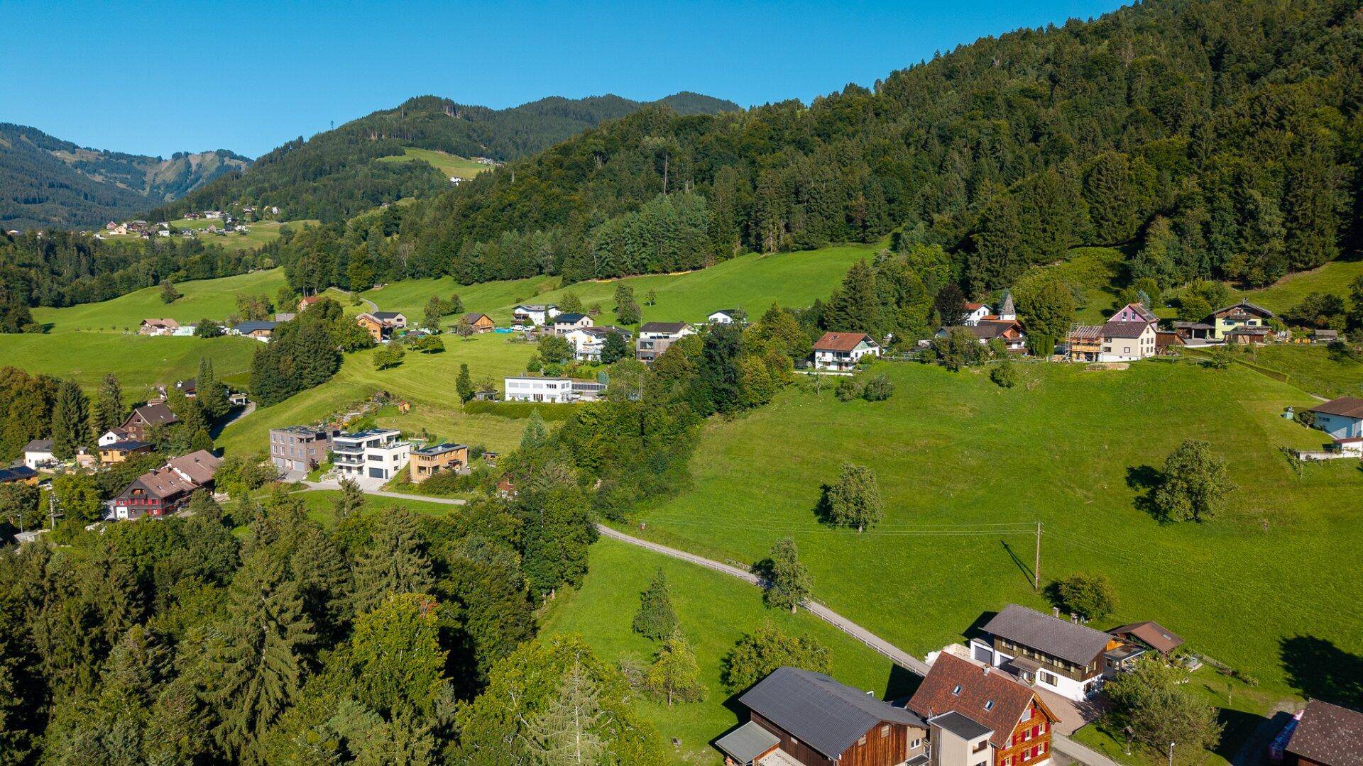 Weitläufige Luftaufnahme eines Dorfes in einer grünen Hügellandschaft mit Bergen im Hintergrund.