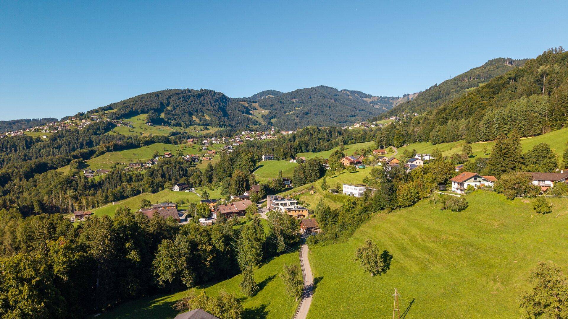 Weitwinkel-Luftaufnahme eines Dorfes in einem grünen Tal mit bewaldeten Hügeln und Bergen.