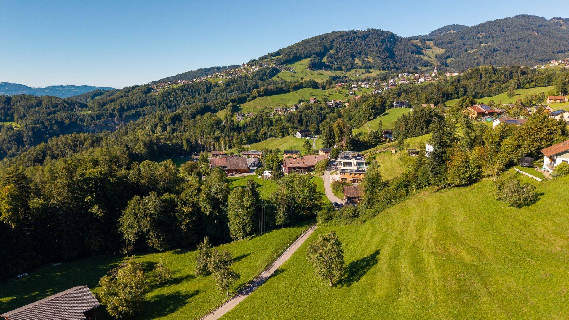 Luftaufnahme eines Dorfes am Hang, umgeben von grünen Feldern und Wäldern mit einer Straße.