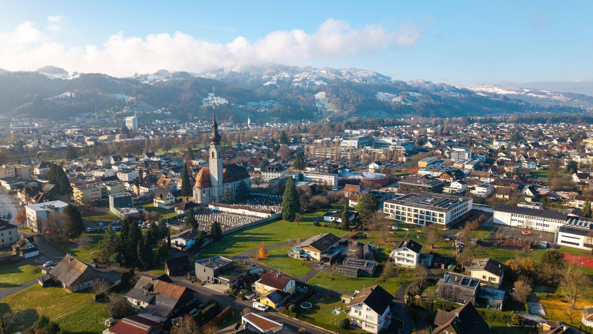 Panoramablick auf eine Stadt mit Kirche, Wohngebieten und schneebedeckten Bergen im Hintergrund.