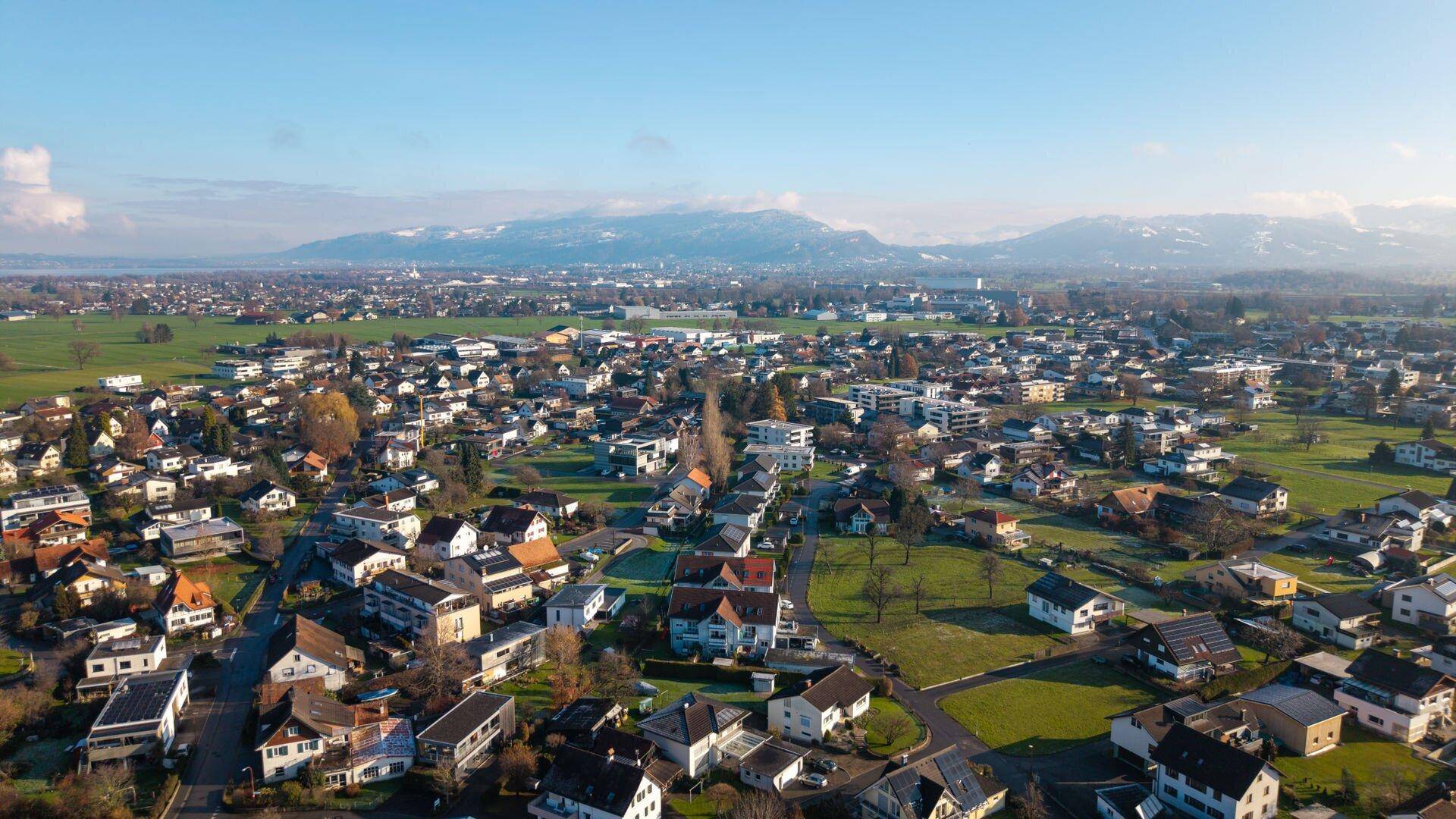 Panoramablick über eine Wohnsiedlung mit grünen Flächen und Bergen am Horizont unter blauem Himmel.