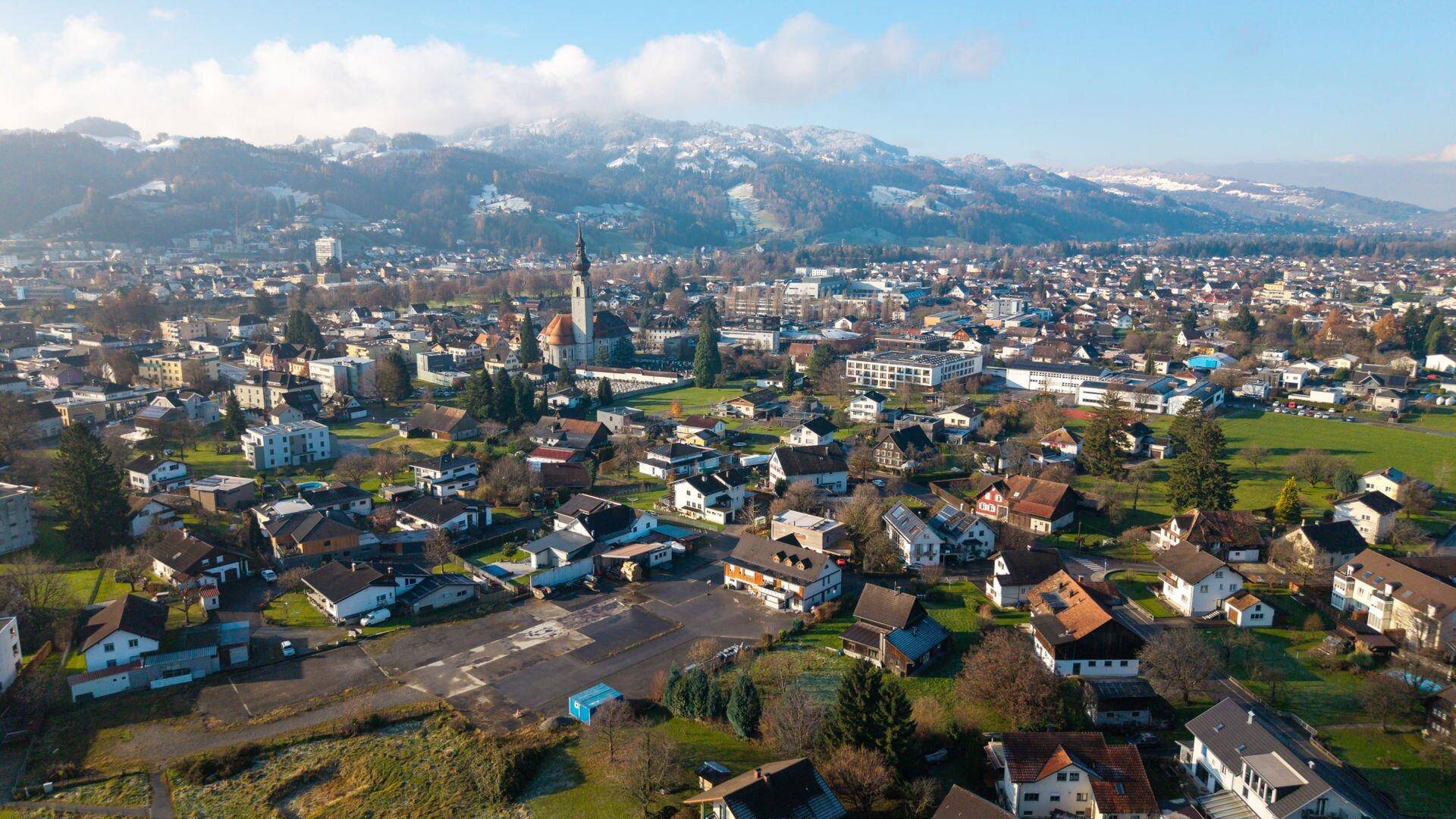 Luftaufnahme einer Stadt mit historischer Kirche, Wohngebieten und schneebedeckten Bergen im Hintergrund.