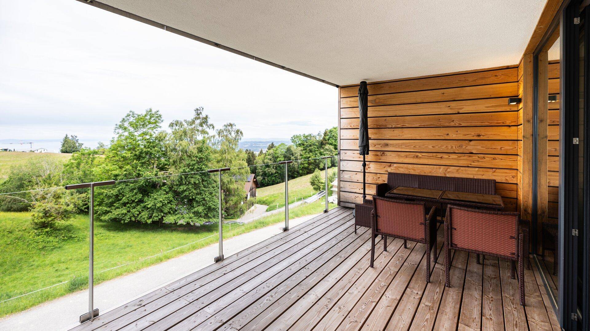 Großer Balkon mit Holzbelag und Glasgeländer, bietet weite Aussicht auf die Landschaft.