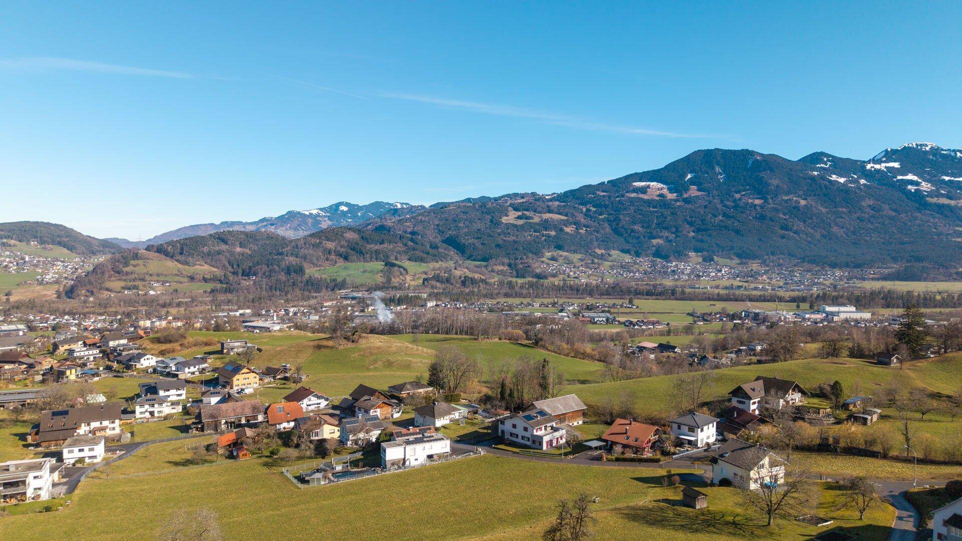 Panoramablick auf eine ländliche Wohnsiedlung mit weiten Grünflächen und einer beeindruckenden Bergkulisse.