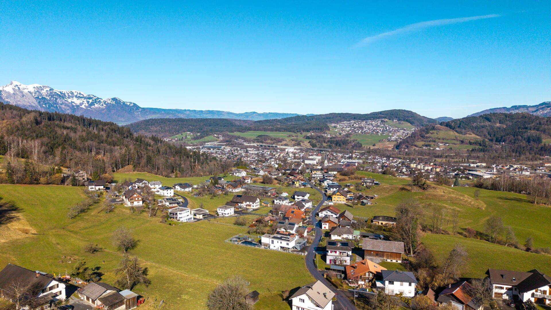Weitläufige Luftaufnahme einer Wohnsiedlung in einer grünen Landschaft mit Bergen und einem weiten Talblick.