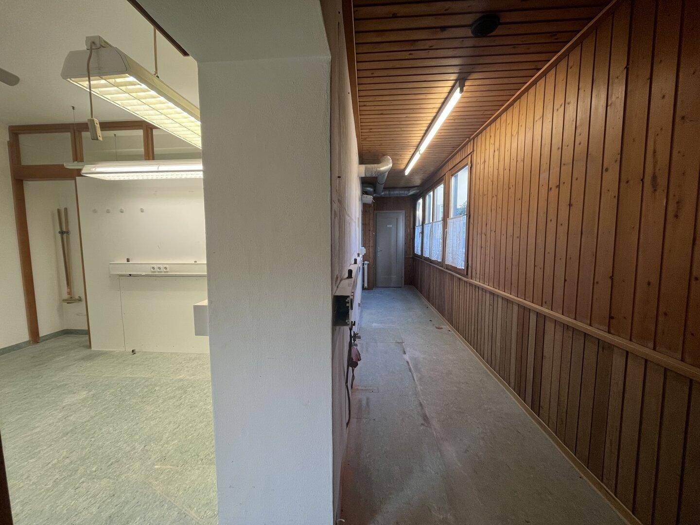 Gangbereich mit Holzvertäfelung an einer Wand und Blick in einen angrenzenden Arbeitsraum.