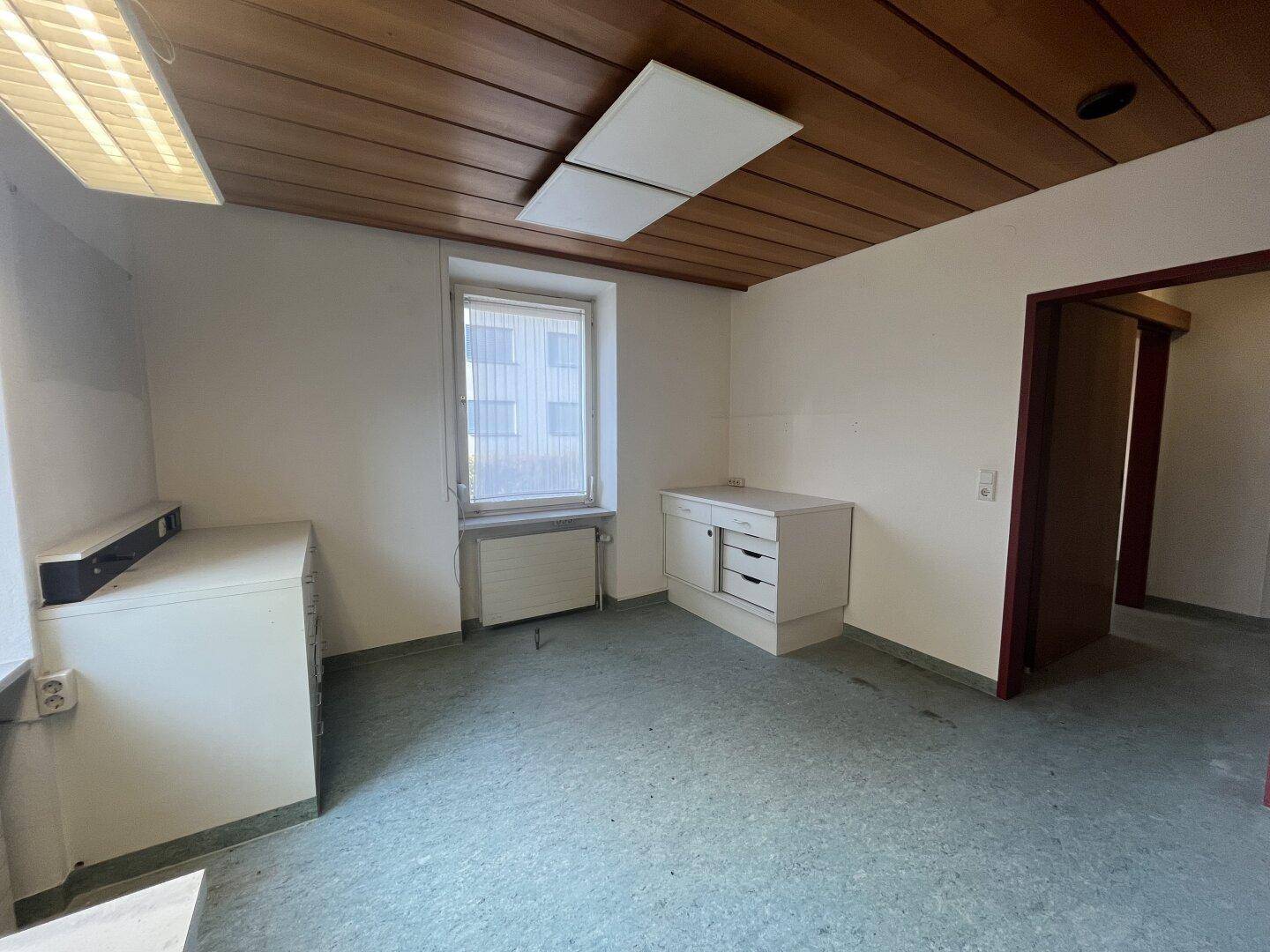 Heller Büroraum mit Holzdecke, Fenster und einfachen weißen Schränken, gut beleuchtet.