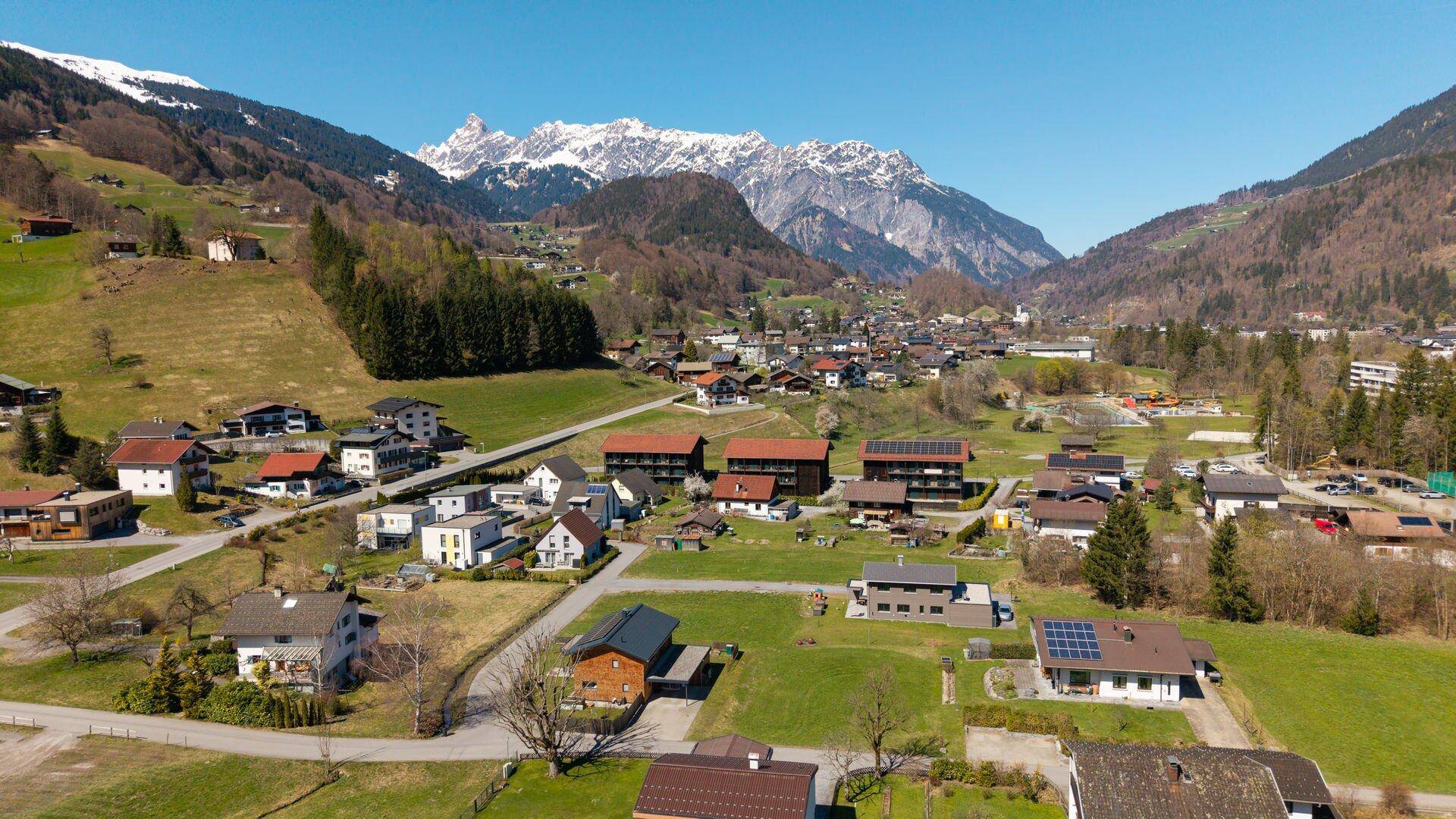 Weitläufige Luftaufnahme eines Dorfes in einem Tal, umgeben von grünen Hügeln und imposanten schneebedeckten Bergen.