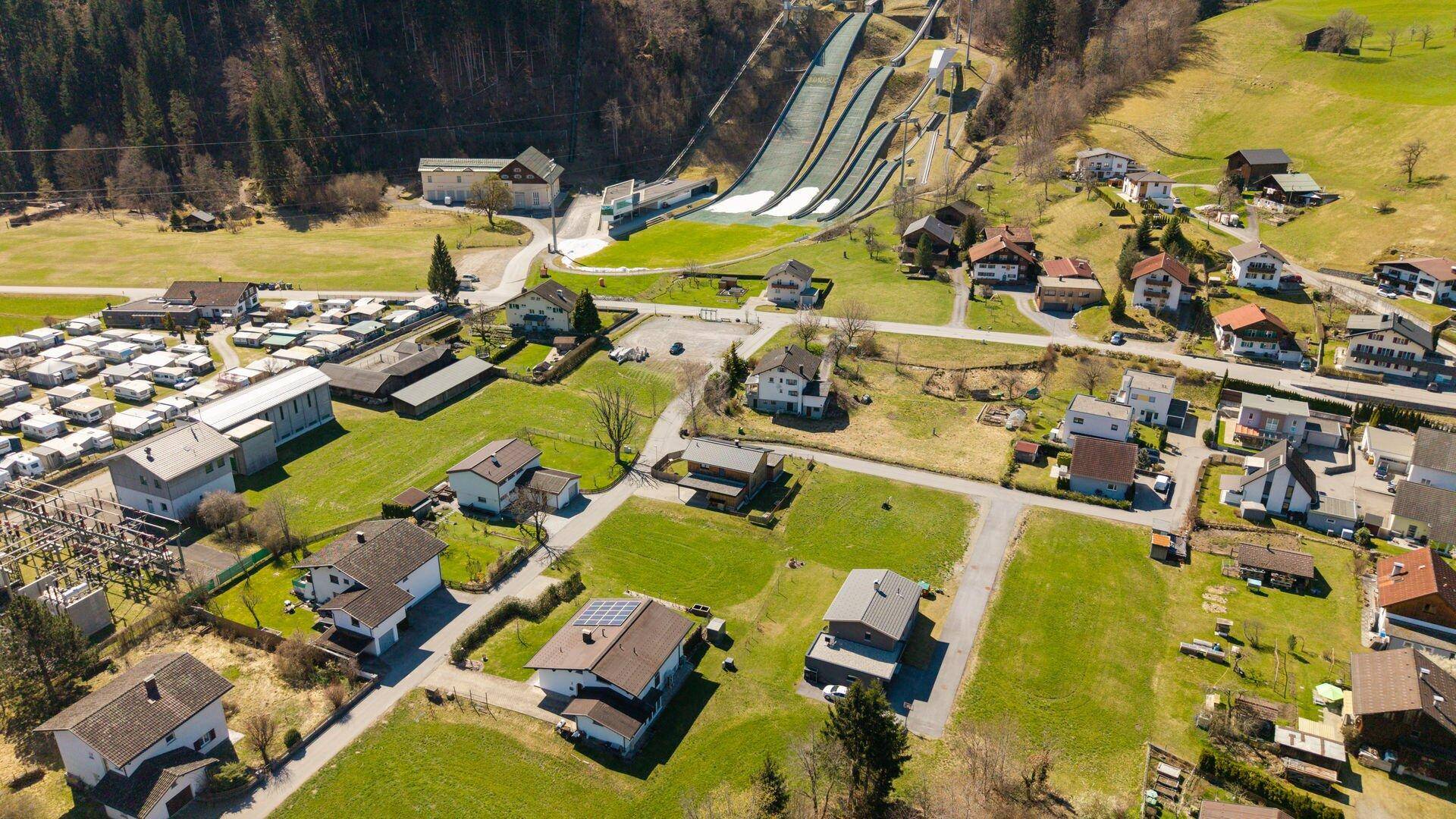 Luftaufnahme einer Wohnsiedlung mit grünen Wiesen und einer Skisprungschanze am Fuße der Berge.