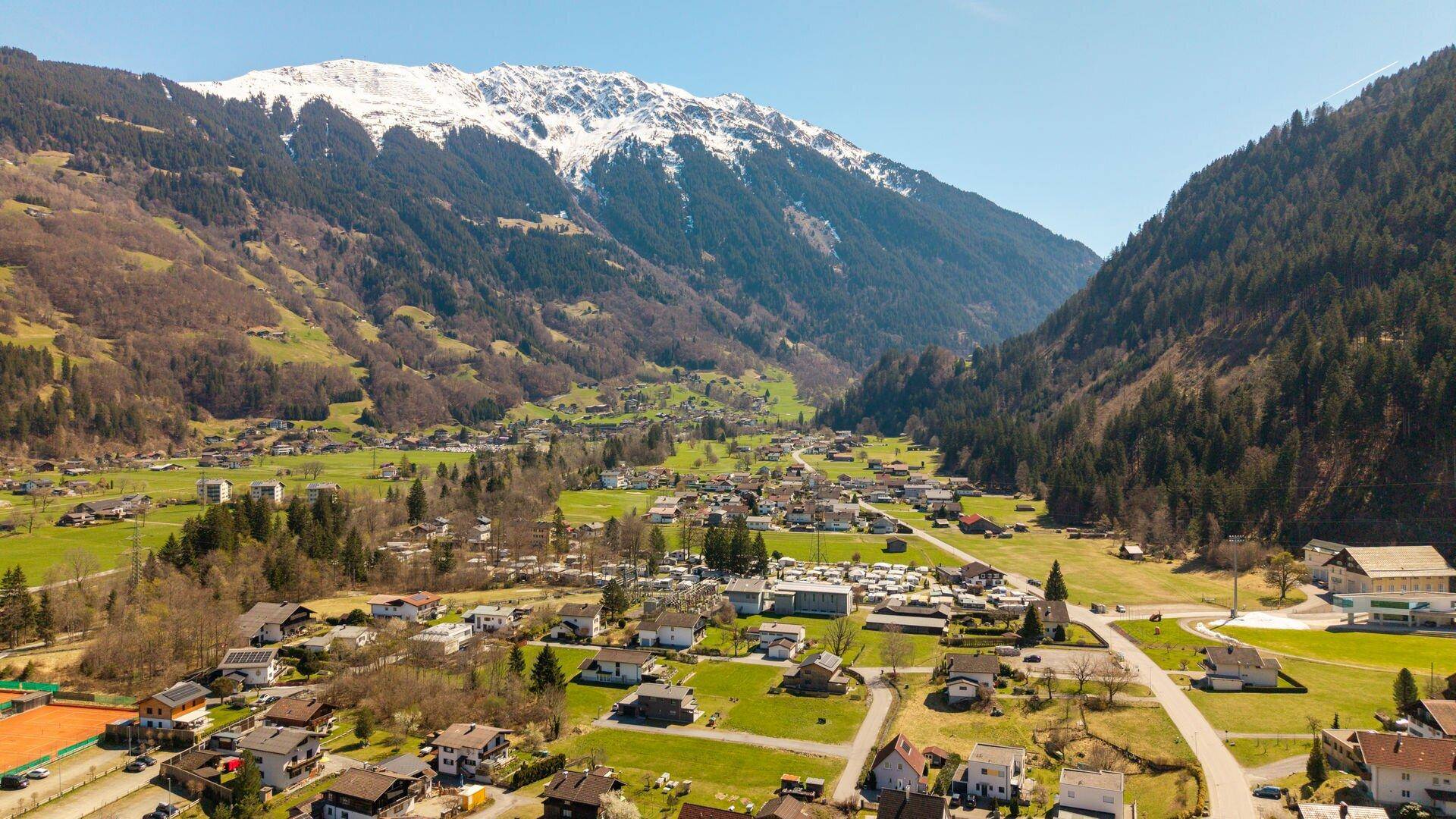 Weitläufige Luftaufnahme eines alpinen Tals mit einer Wohnsiedlung, umgeben von schneebedeckten Bergen und Wäldern.