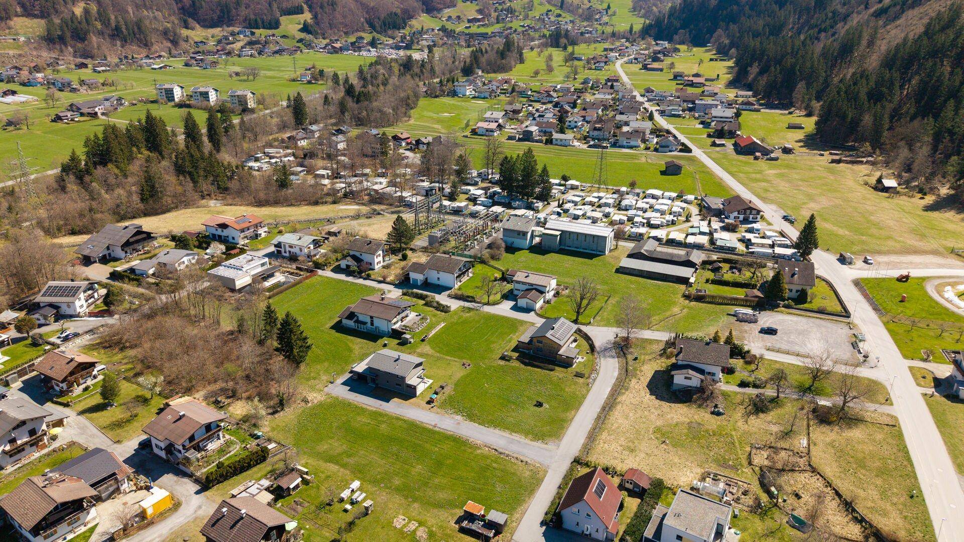Luftaufnahme einer Wohnsiedlung in einem weiten Tal mit grünen Feldern und einer Bergkulisse im Hintergrund.