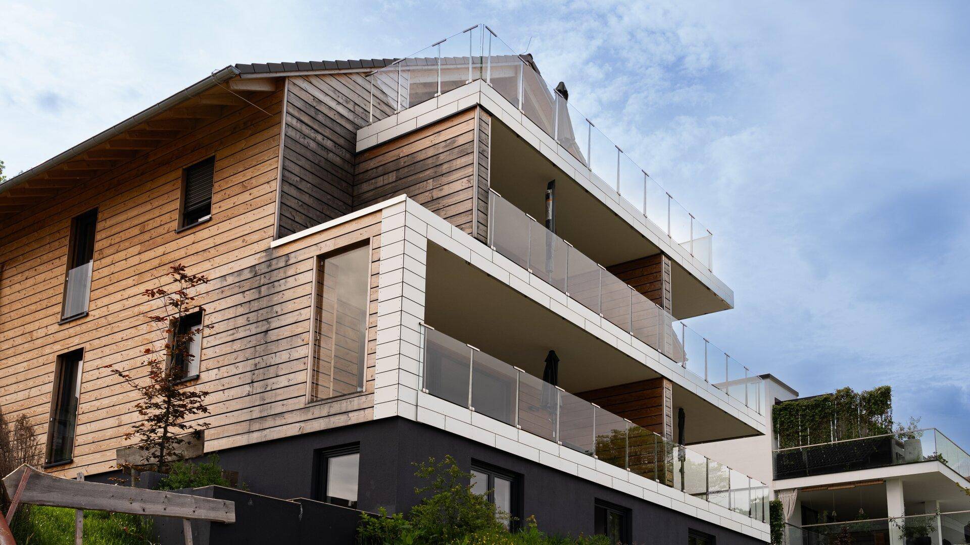 Moderne Architektur mit Holzfassade und mehreren Balkonen mit Glasgeländer unter bewölktem Himmel.