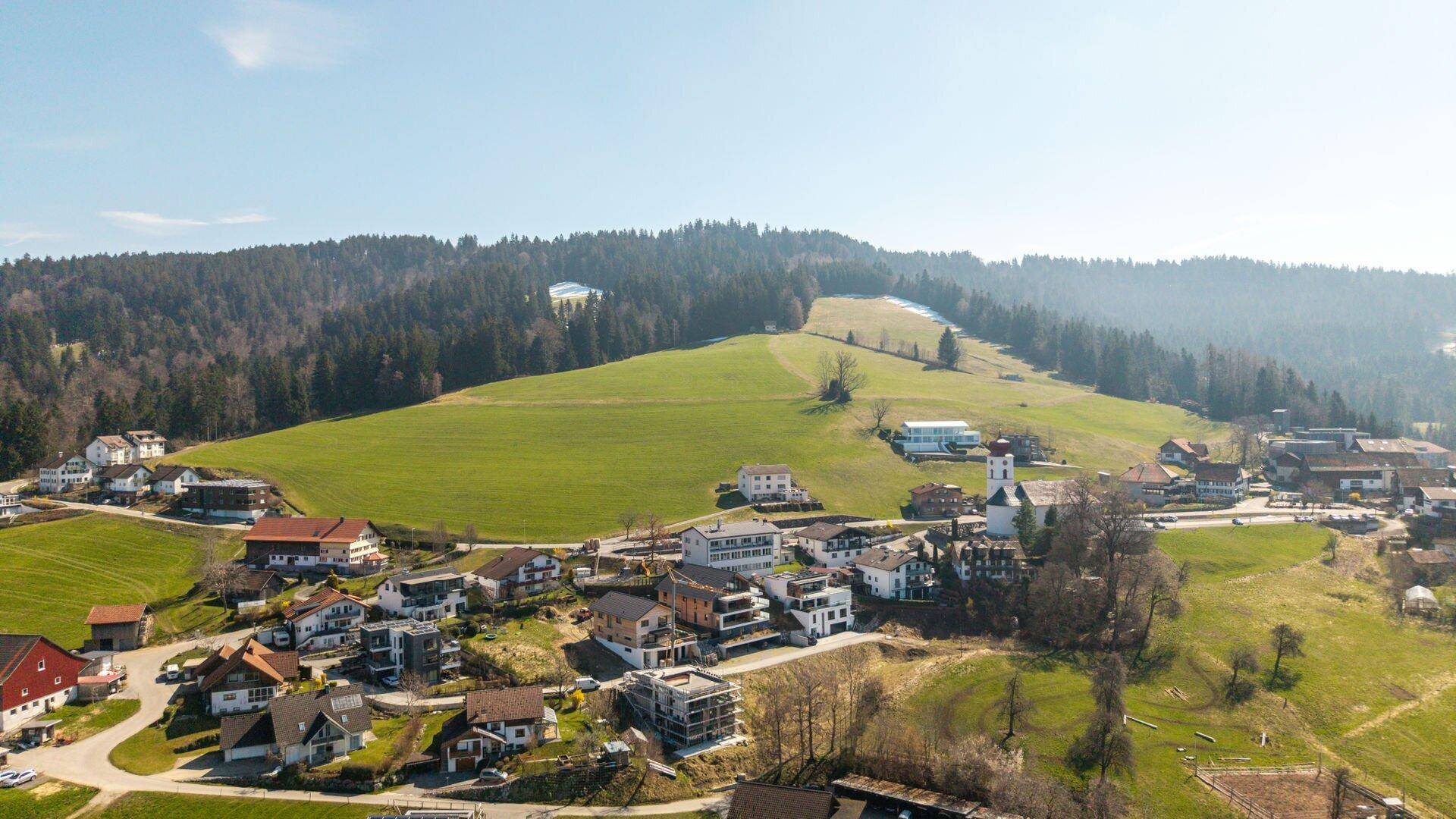 Luftaufnahme eines malerischen Dorfes eingebettet in eine grüne Hügellandschaft mit Wald.