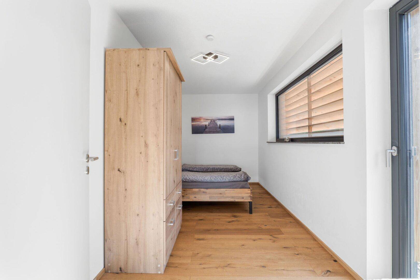 Helles Schlafzimmer mit Holzbett, Kleiderschrank und Fenster mit Jalousien.