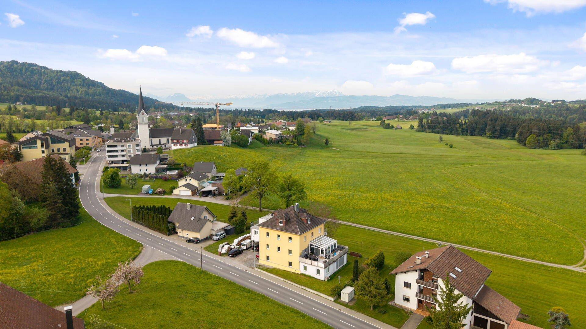 Luftaufnahme einer Ortschaft mit Kirche, grünen Feldern und Bergen im Hintergrund.