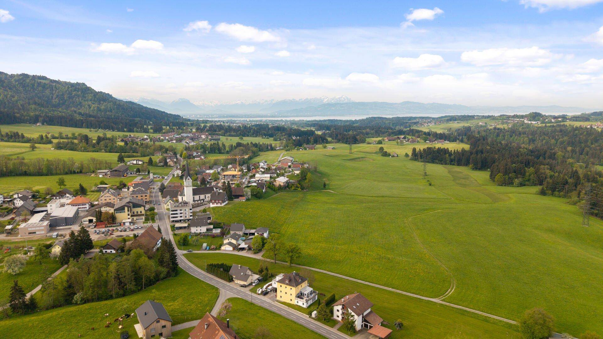 Panoramablick aus der Luft auf eine Ortschaft, grüne Landschaft und einen See in der Ferne.
