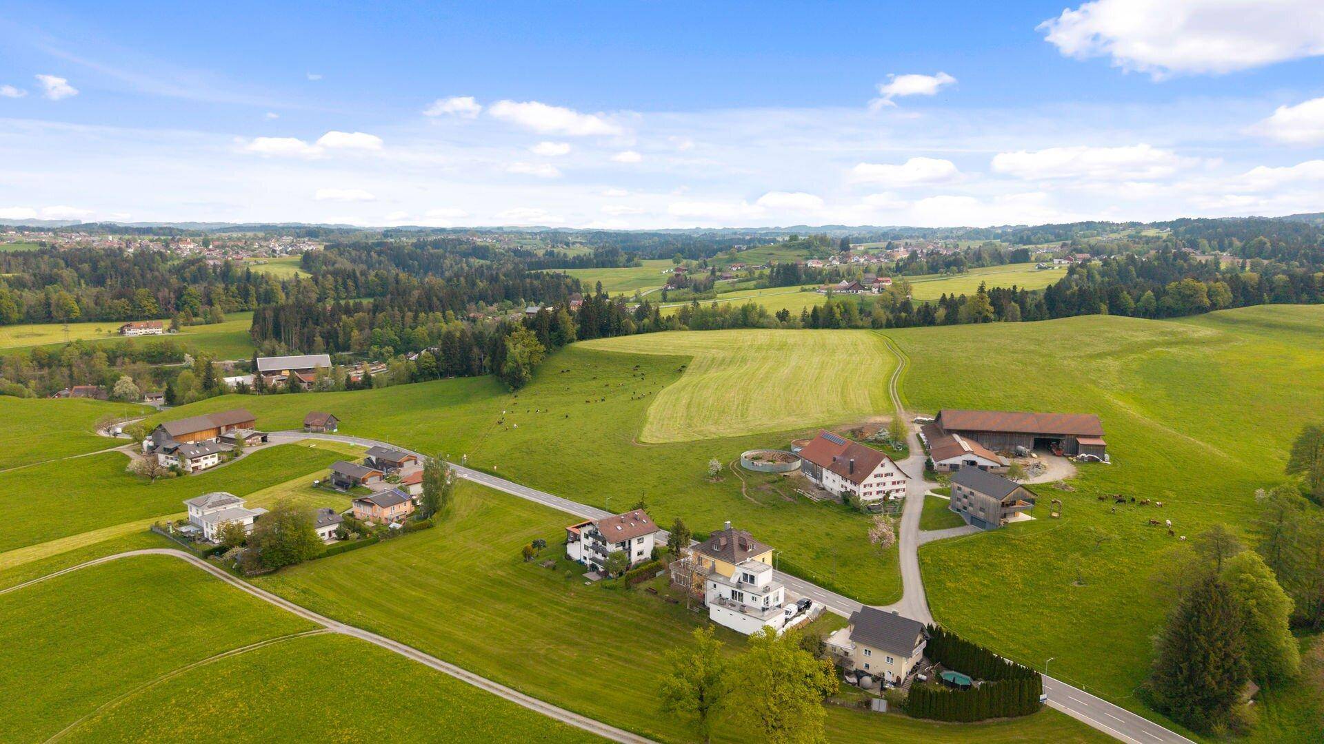 Luftaufnahme einer ländlichen Siedlung, umgeben von weiten grünen Wiesen und Wäldern.