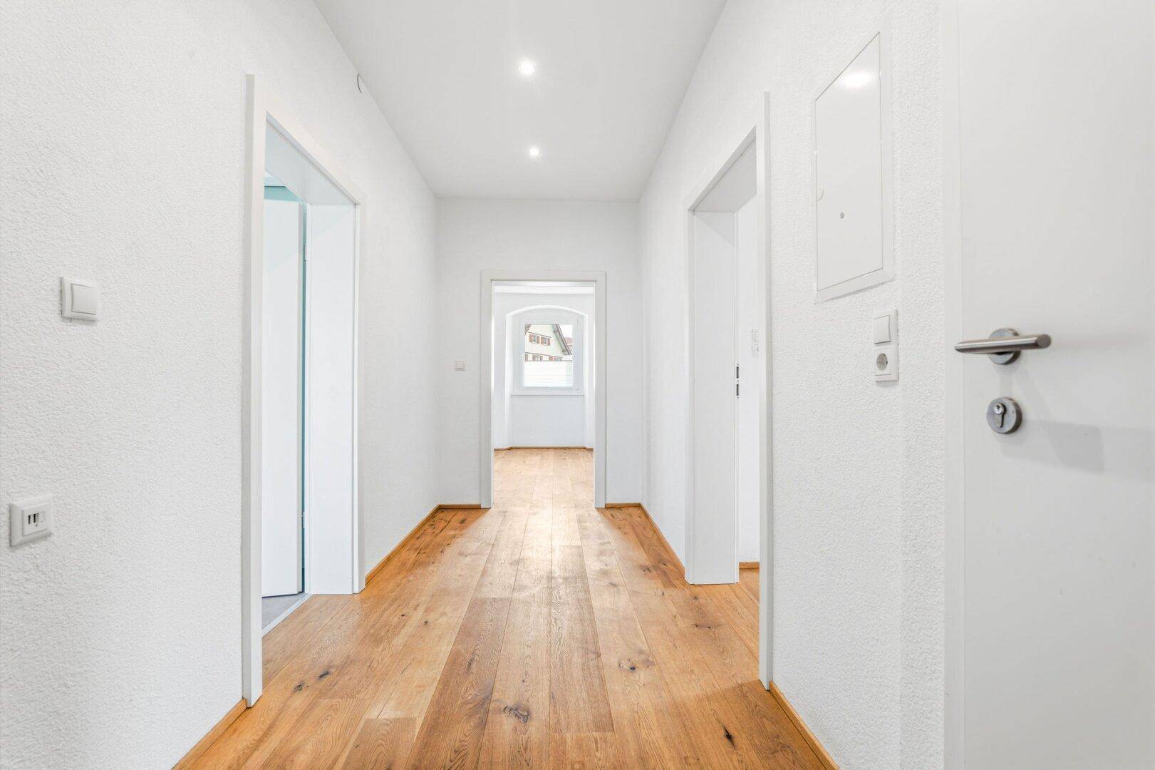Langer Flur mit Holzdielenboden, weißen Wänden und Blick in einen weiteren Raum.