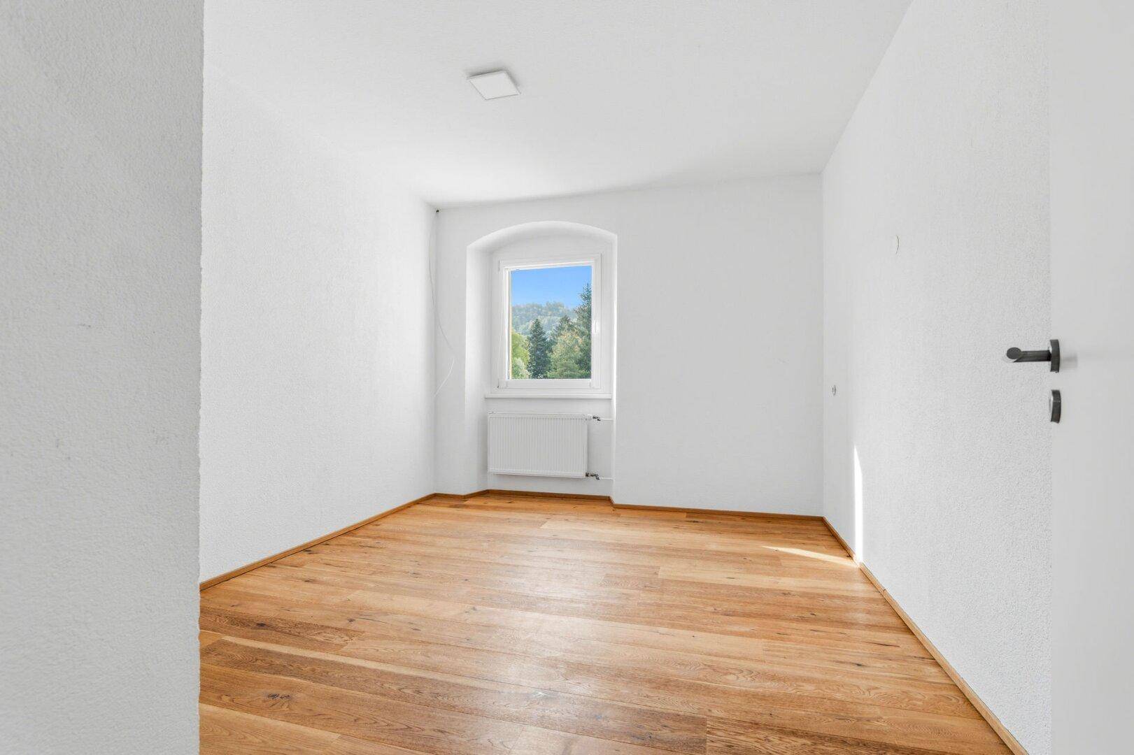 Helles Schlafzimmer mit Holzdielenboden, weiß gestrichenen Wänden und Fenster mit Grünblick.