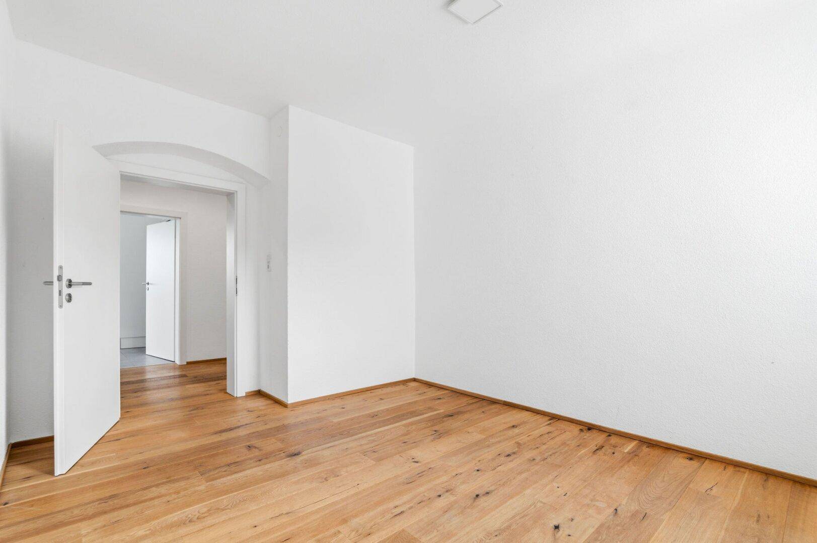 Heller Wohnraum mit Holzdielenboden und weiß gestrichenen Wänden, offen zu weiteren Räumen.