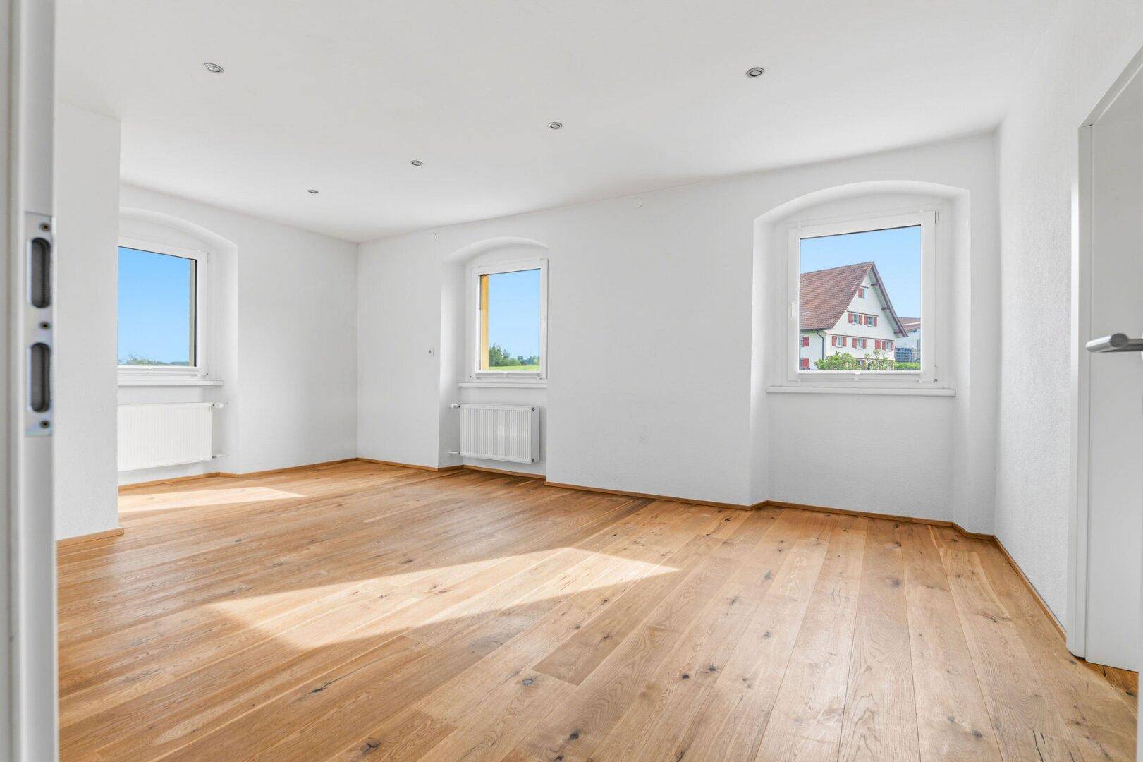 Lichtdurchfluteter Wohnraum mit Holzdielenboden und mehreren Fenstern mit Blick ins Grüne.