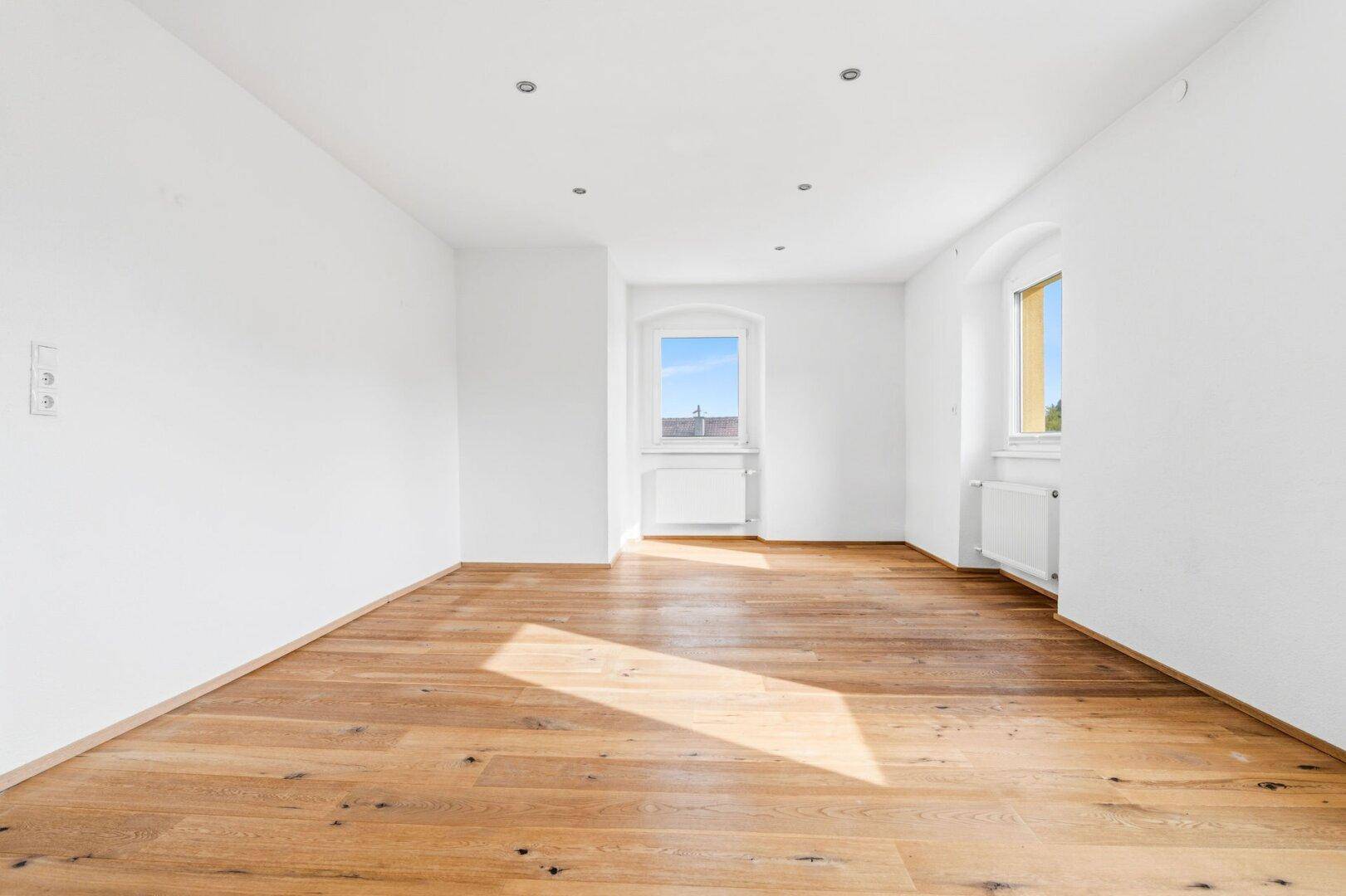 Geräumiger Wohnbereich mit Holzdielenboden, weißen Wänden und Einbaustrahlern an der Decke.