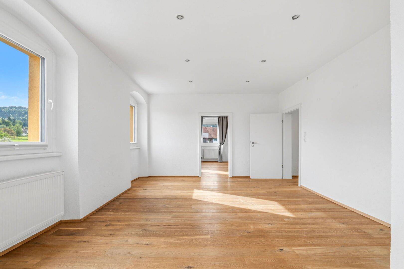 Heller Wohnraum mit Holzdielenboden, weißen Wänden und Zugang zu weiteren Zimmern.