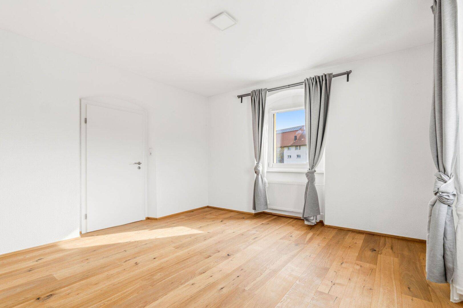 Geräumiges Schlafzimmer mit Holzdielenboden, weißen Wänden und zwei Fenstern mit Vorhängen.