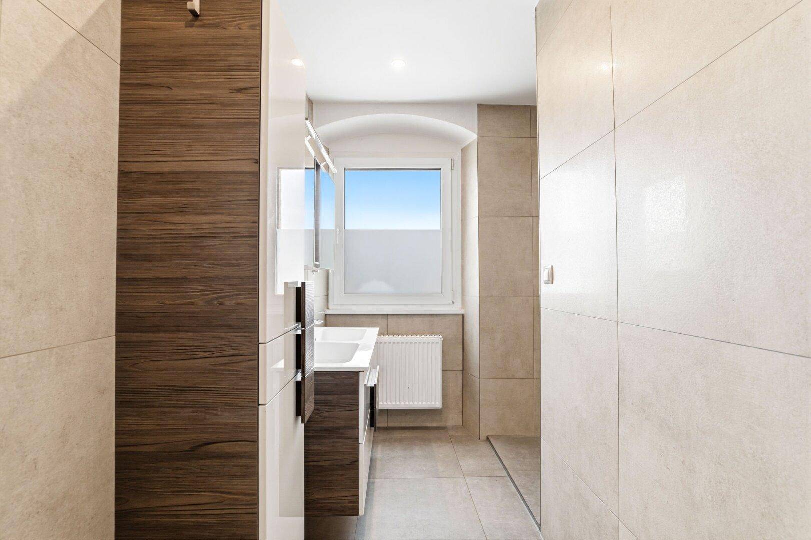 Modernes Badezimmer mit beigefarbenen Fliesen, Waschtisch und Fenster für Tageslicht.