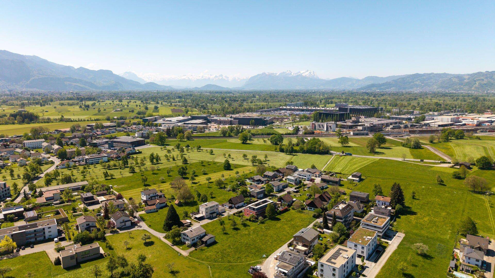 Luftaufnahme einer Siedlung mit weiten Grünflächen und einer beeindruckenden Bergkette in der Ferne.