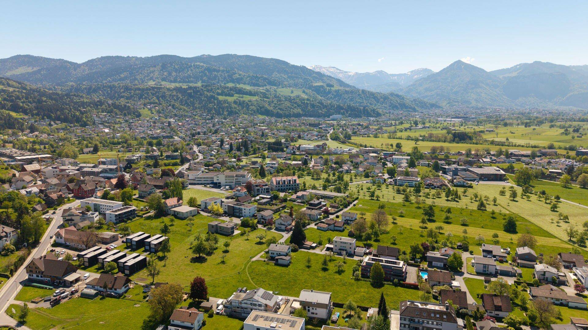 Panoramische Luftansicht einer Wohnsiedlung, umgeben von grünen Hügeln und schneebedeckten Bergen.