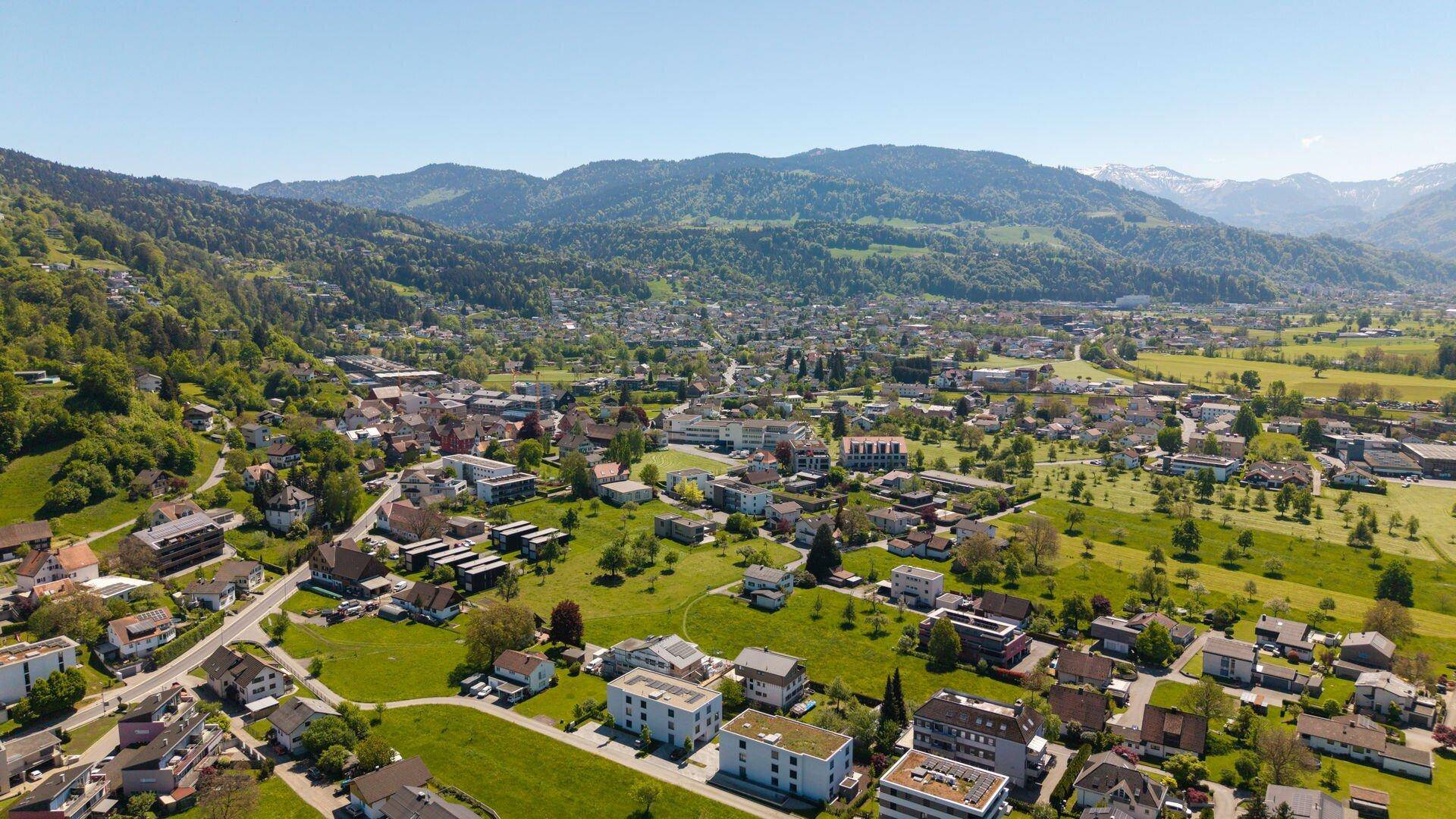 Luftaufnahme einer weitläufigen Wohngegend mit grünen Wiesen und einer malerischen Bergkulisse im Hintergrund.