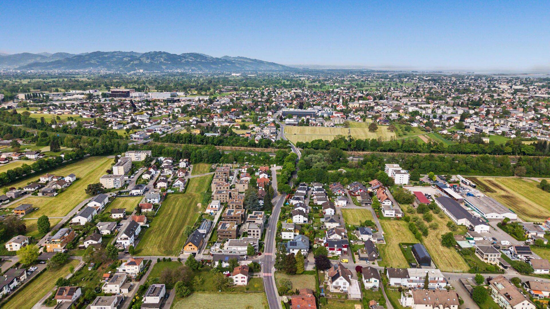 Panoramische Luftaufnahme einer Siedlung mit Wohngebäuden, Feldern und einer Bergkette am Horizont.
