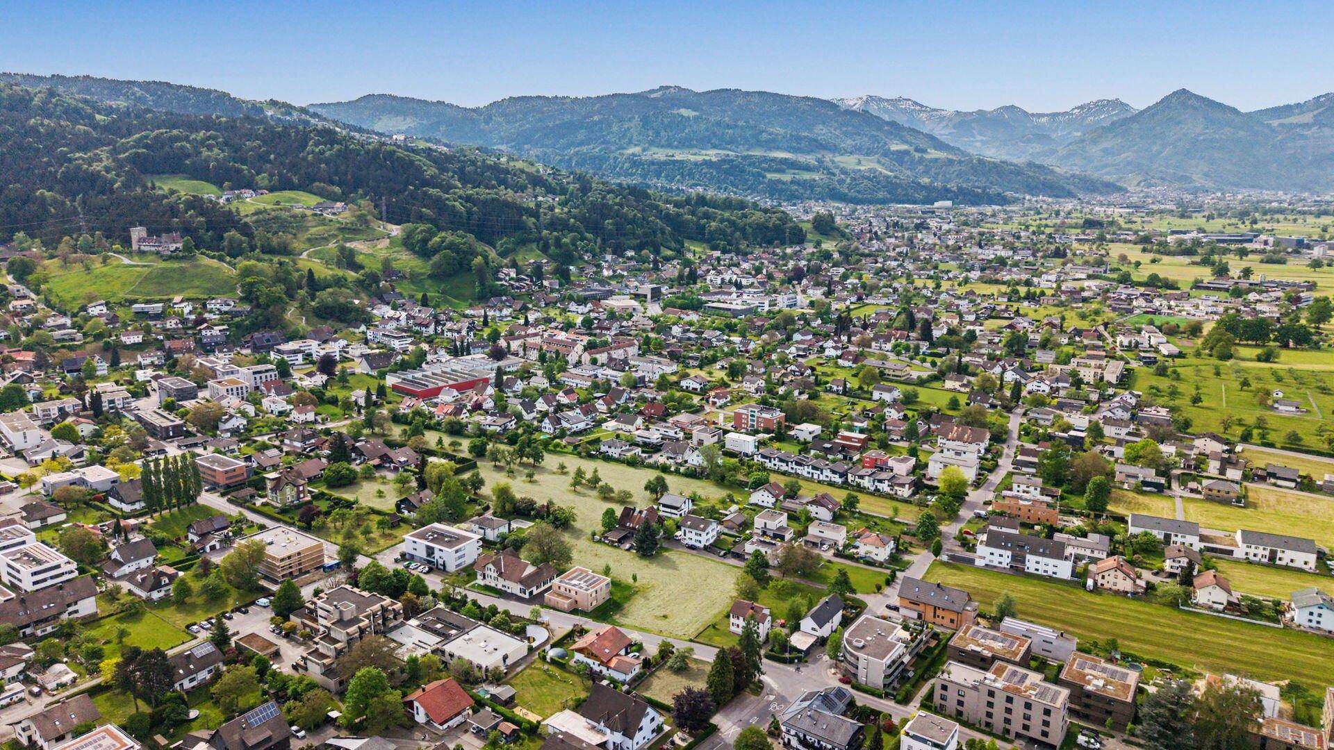 Luftaufnahme einer Wohnsiedlung mit grünen Hügeln und Bergen im Hintergrund unter blauem Himmel.