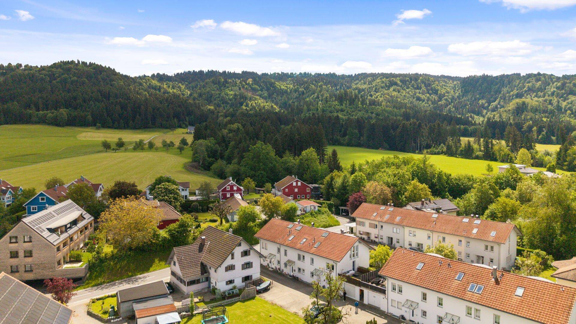 Weitläufige Luftaufnahme einer ländlichen Siedlung umgeben von grünen Hügeln und Wäldern.
