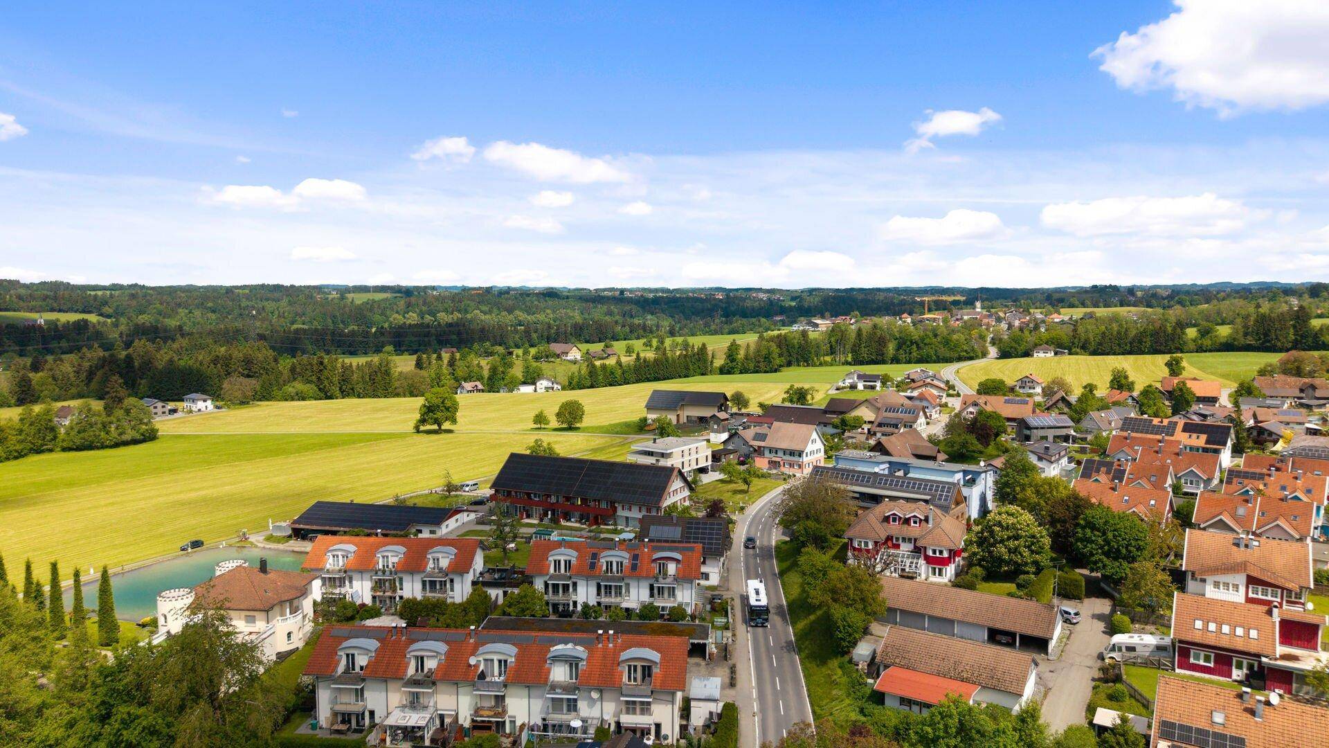 Luftaufnahme einer Wohnsiedlung mit einer Straße, umgeben von Feldern und Wäldern unter blauem Himmel.